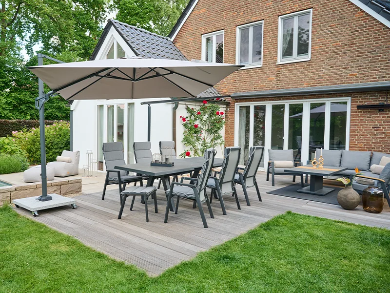 Garten-Esstischgarnitur mit hellen Stühlen unter großem Ampelschirm auf Holzterrasse vor moderner Hausfront mit großen Fensterflächen.